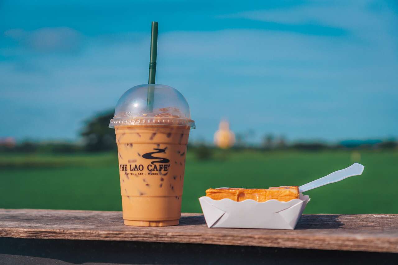 Iced Lao Coffee on a wooden table by a rice field with the Great Buddha of Thailand in the background.