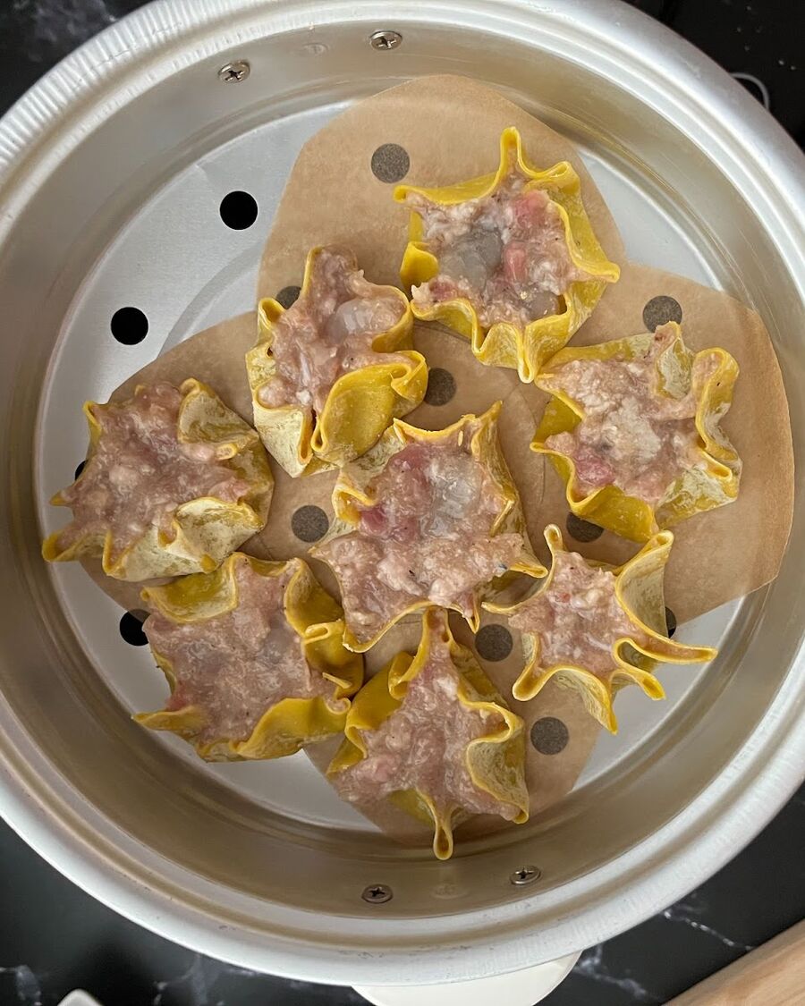 Pour 1-2 cups of water into the rice cooker. Utilize the steaming basket provided with the cooker. Place a cooking paper sheet over the basket, ensuring to leave holes for steam release. Activate the cooker and steam the dumplings for approximately 15 minutes, checking periodically.