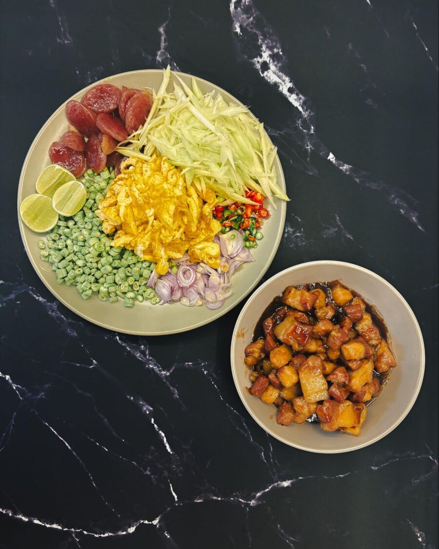 Prepare all the condiments: shred the green mango, slice the shallots, chop the long beans and fresh chilies, slice the omelet into strips, cut fresh lime wedges, and slice the Chinese sausage and caramelized pork.